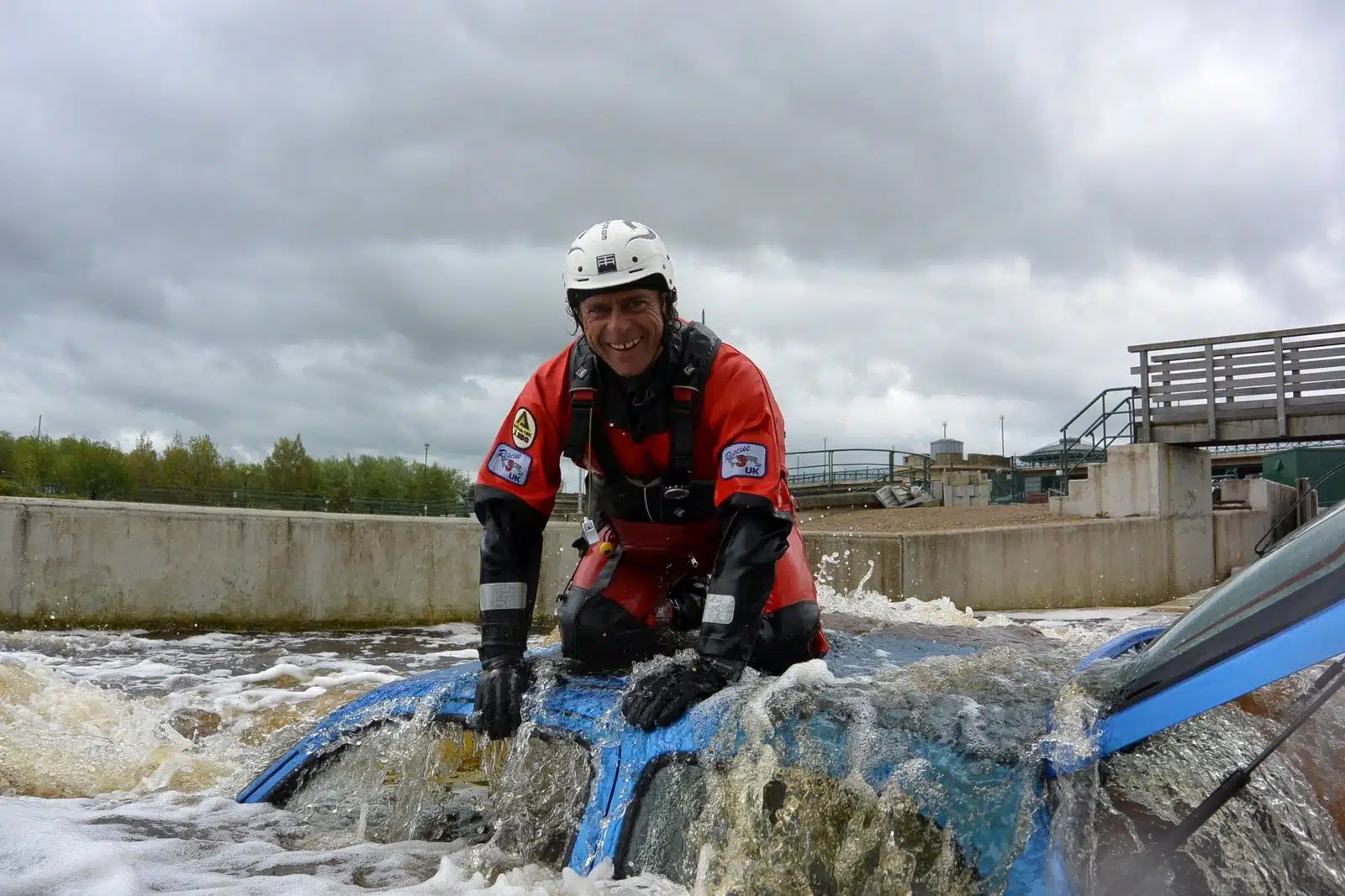 Soccorso Alluvionale e Fluviale Europa Un soccorritore, che ricorda una scena di David Alemanni, è in piedi, in piena attrezzatura, in cima a un'auto parzialmente sommersa, mentre lotta contro l'acqua impetuosa sotto un cielo coperto. S.A.F.E.® - River Rescue