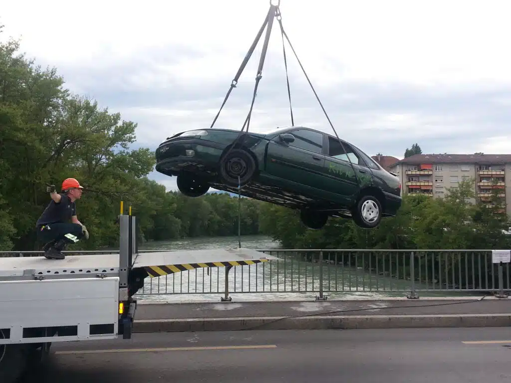 Soccorso Alluvionale e Fluviale Europa Un'auto verde viene sollevata da una gru su un ponte fluviale, ricordando una scena di Rescue 3, con un operaio che la guida con perizia su un camion a pianale, dimostrando la competenza nel soccorso ad autoveicoli immersi nel fiume. S.A.F.E.® - River Rescue