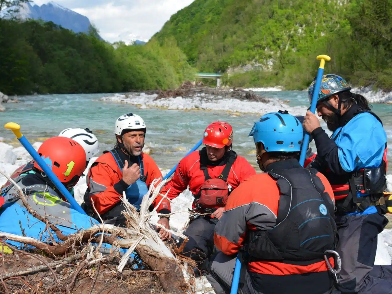 Soccorso Alluvionale e Fluviale Europa Un gruppo di persone con casco e attrezzatura, guidate da un istruttore esperto, discutono in riva a un fiume circondato da montagne e vegetazione, apparentemente in preparazione per un'operazione di soccorso di terza classe. S.A.F.E.&reg; - River Rescue