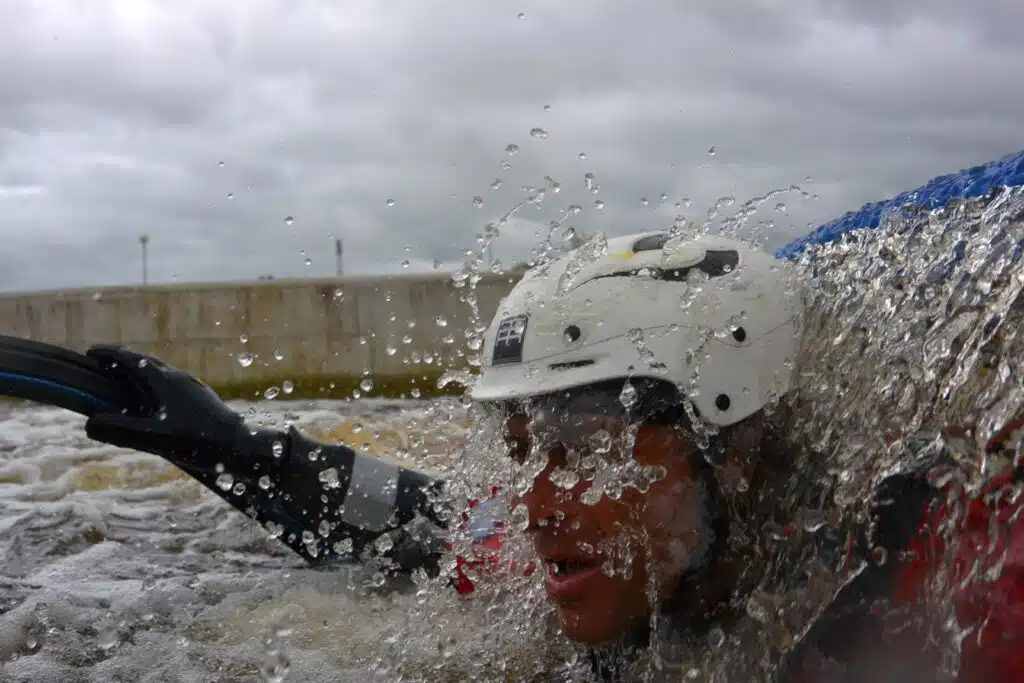 Soccorso Alluvionale e Fluviale Europa Una persona con il casco, simbolo di un'operazione Rescue 3, è parzialmente immersa nell'acqua, circondata da schizzi, sotto un cielo nuvoloso. S.A.F.E.® - River Rescue