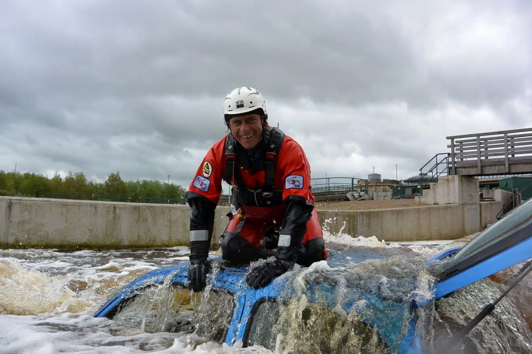 Soccorso Alluvionale e Fluviale Europa Un operatore di Rescue 3 in tenuta completa si inginocchia su un'auto sommersa, occupandosi con perizia del soccorso ad autoveicoli immersi nel fiume, il tutto sotto la distesa vigile di un cielo nuvoloso. S.A.F.E.® - River Rescue