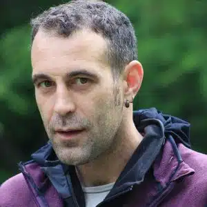 Soccorso Alluvionale e Fluviale Europa Filippo Villa, con i capelli corti e un orecchino a cerchio, indossa una giacca viola con cappuccio nero mentre è in piedi in mezzo a una vegetazione lussureggiante. S.A.F.E.® - River Rescue