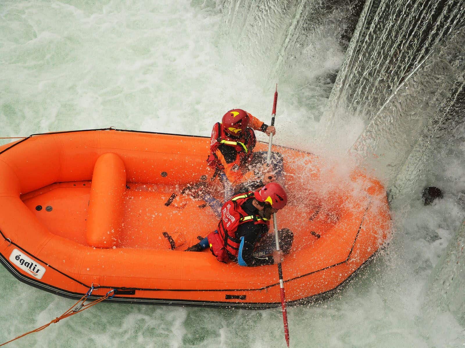 Flood and River Rescue Europe Two people in an orange inflatable boat skilfully negotiate the swift and turbulent waters near a waterfall, equipped with Rescue 3 helmets and life jackets. S.A.F.E.® - River Rescue