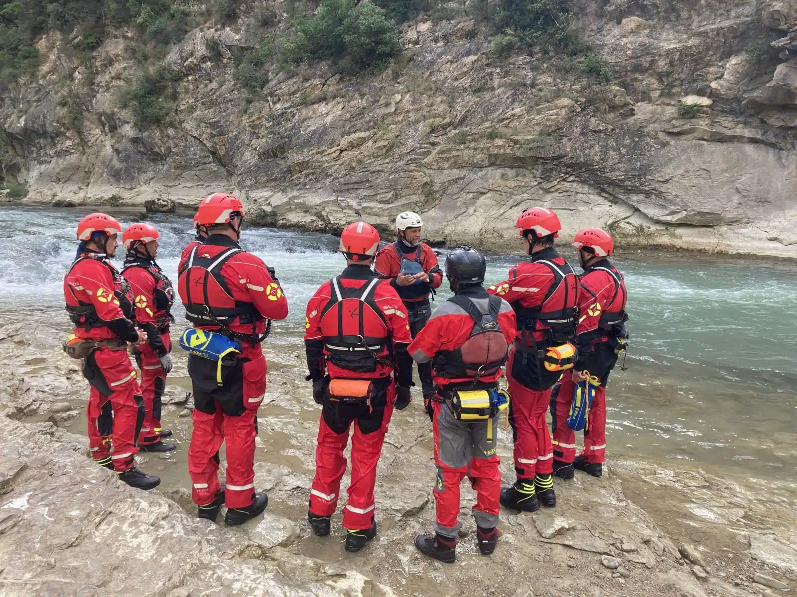 Soccorso Alluvionale e Fluviale Europa La squadra di soccorso 3, in tenuta rossa, &egrave; in piedi su una riva rocciosa del fiume e discute la strategia nei pressi di un fiume che scorre rapidamente. S.A.F.E.&reg; - River Rescue