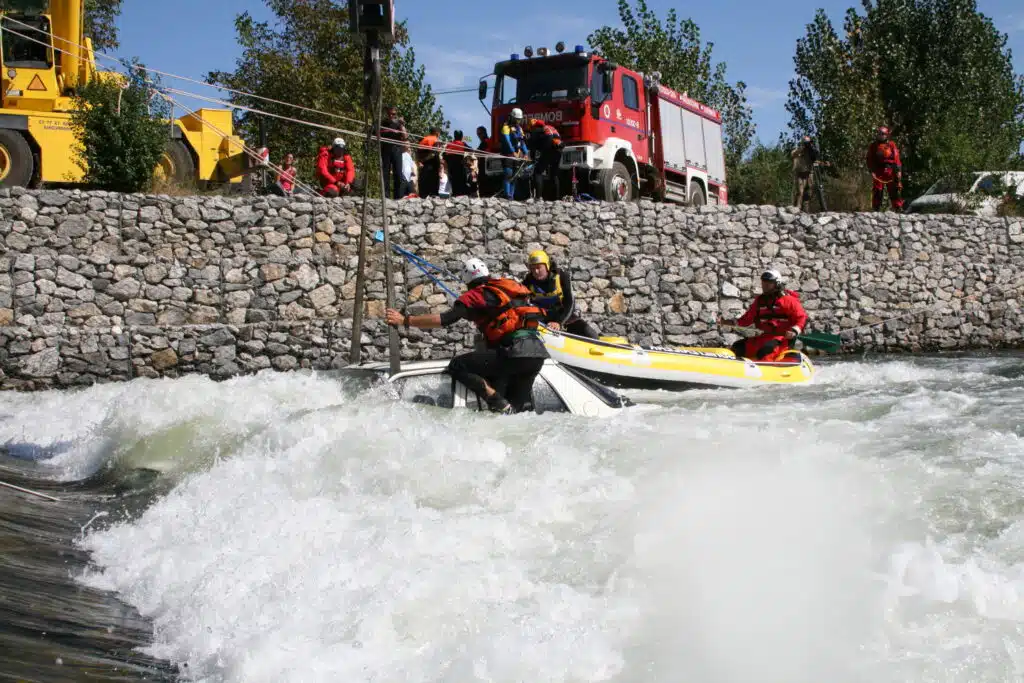 Soccorso Alluvionale e Fluviale Europa I soccorritori a bordo di una zattera prestano soccorso a un'imbarcazione ribaltata in una rapida vicino a un muro di pietra, con spettatori e un camion di soccorso nelle vicinanze. S.A.F.E.® - River Rescue
