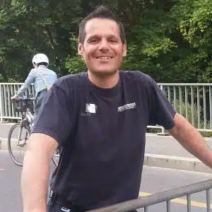 Soccorso Alluvionale e Fluviale Europa Un uomo sorridente con una camicia blu navy, forse Nicolas Millot, è in piedi su un ponte, con un ciclista che si muove sullo sfondo. S.A.F.E.® - River Rescue
