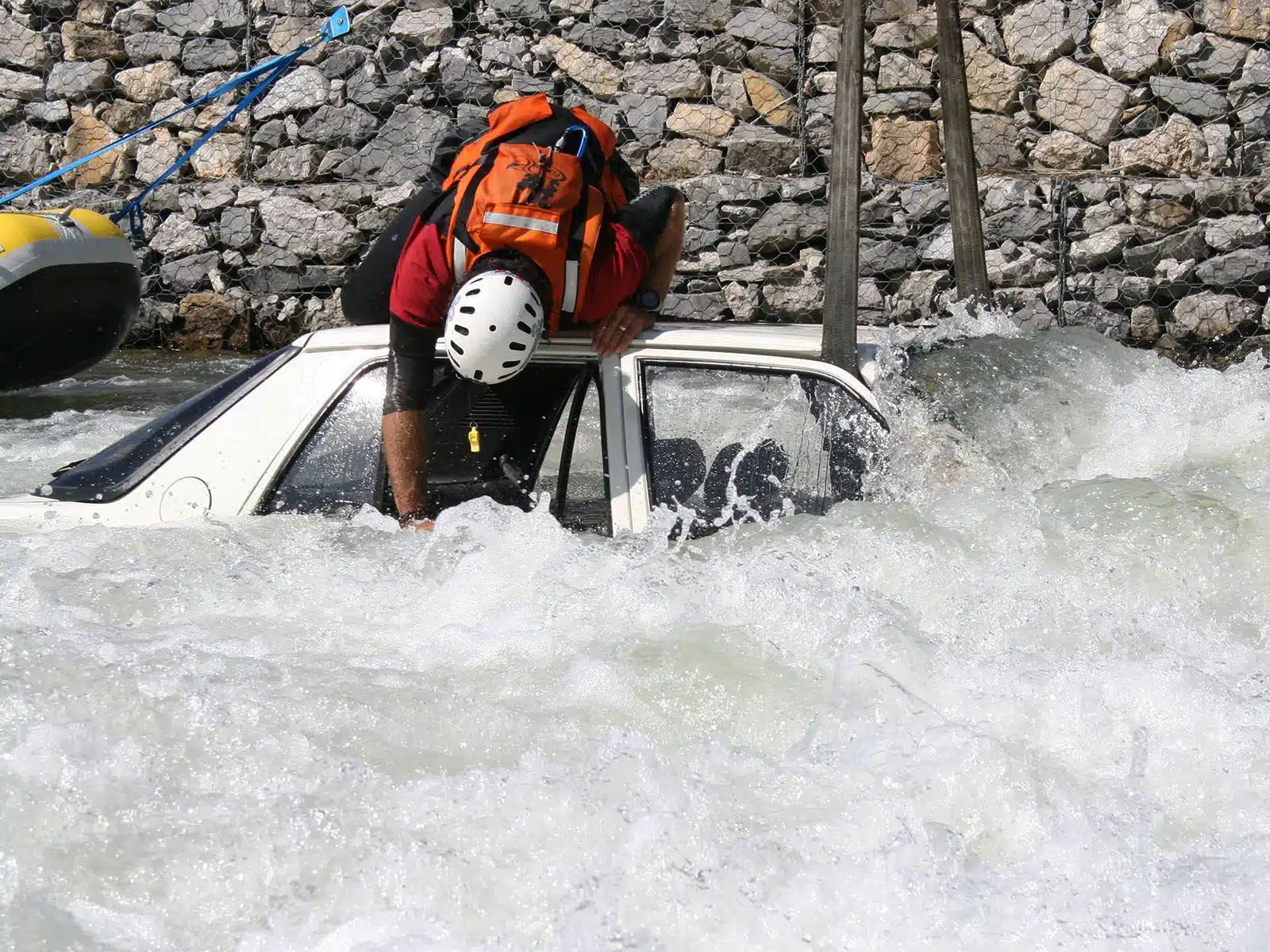 Soccorso Alluvionale e Fluviale Europa Un membro del soccorso 3 si sporge dentro un'auto bianca sommersa dall'acqua corrente vicino a un muro di pietra, con il casco che brilla sotto la luce del sole. S.A.F.E.&reg; - River Rescue