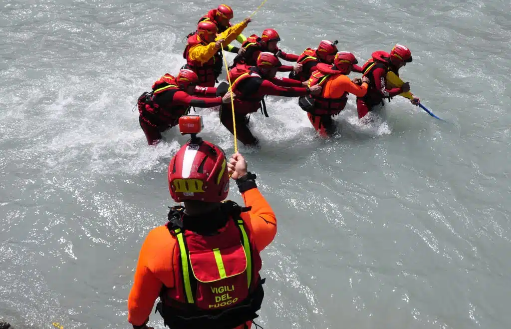 Soccorso Alluvionale e Fluviale Europa La squadra del Soccorso Fluviale di Rovereto, vestita con vivaci tenute rosse e gialle, attraversa il fiume con precisione. Il loro capo naviga abilmente la corrente rapida, afferrando una corda mentre tutti seguono la sua guida. S.A.F.E.® - River Rescue