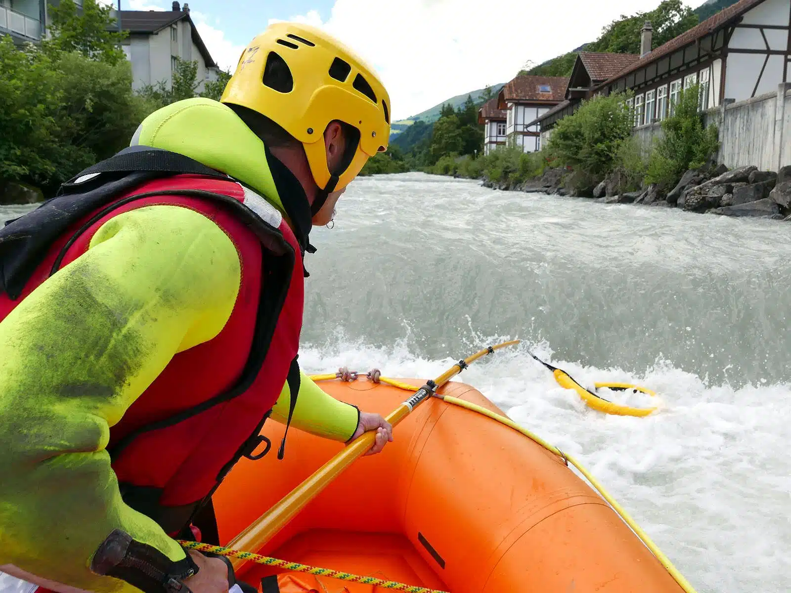 Soccorso Alluvionale e Fluviale Europa Una persona con casco e giubbotto salvagente naviga nel fiume in tempesta con la competenza di un professionista del Rescue 3, circondata da una vegetazione lussureggiante e da edifici sparsi. S.A.F.E.&reg; - River Rescue