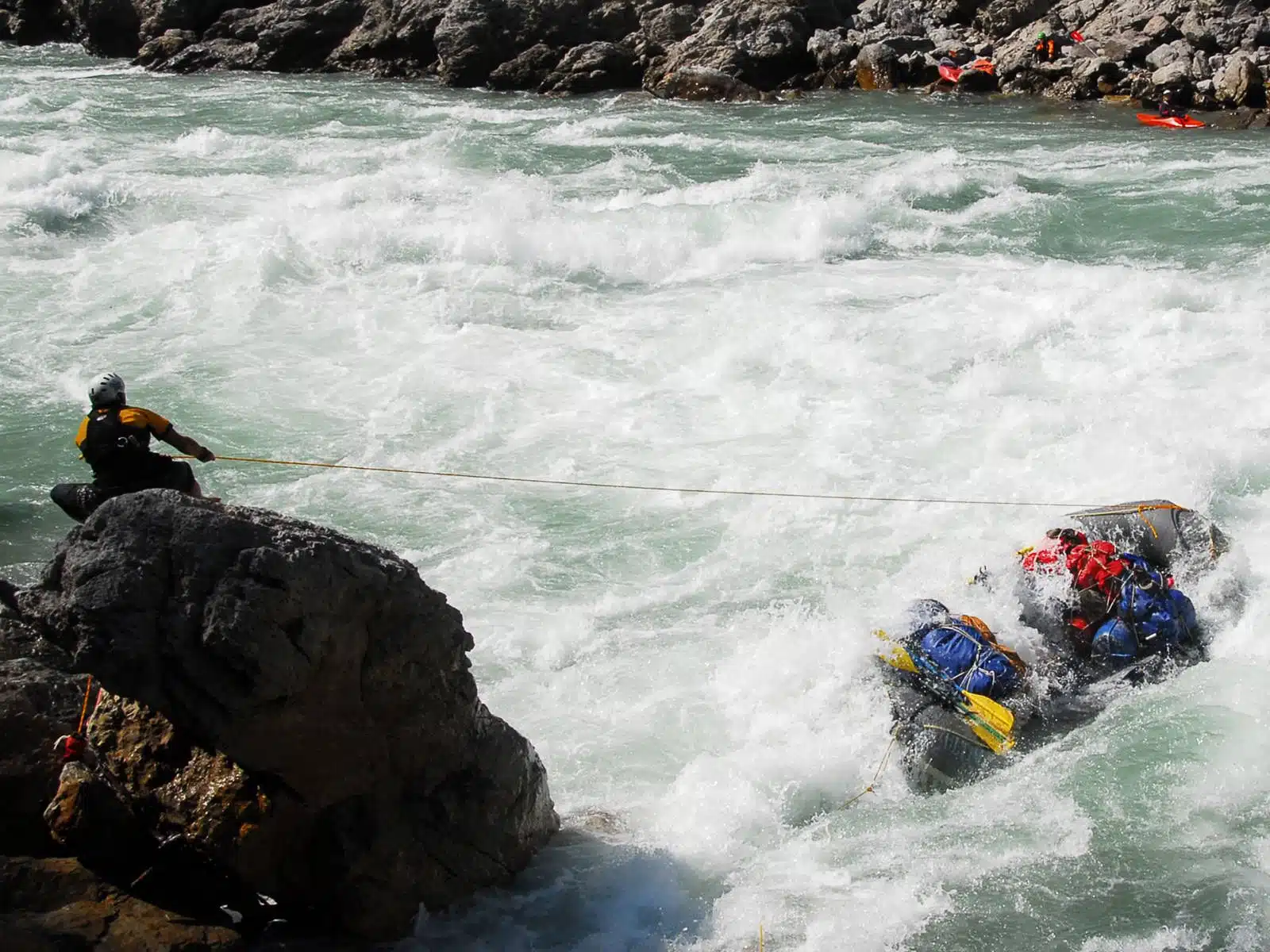 Soccorso Alluvionale e Fluviale Europa In mezzo alle rapide impetuose, un uomo intraprende coraggiosamente un'intensa operazione di salvataggio, manovrando abilmente una zattera piena di persone attraverso le tumultuose acque bianche, il tutto mentre naviga su un difficile terreno roccioso con nient'altro che una robusta corda. S.A.F.E.&reg; - River Rescue