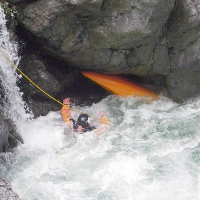 Flood and River Rescue Europe A person wearing a helmet and orange kayak, probably a kayak guide, gets trapped between rocks in turbulent water, clinging to a rope near a small waterfall. S.A.F.E.® - River Rescue