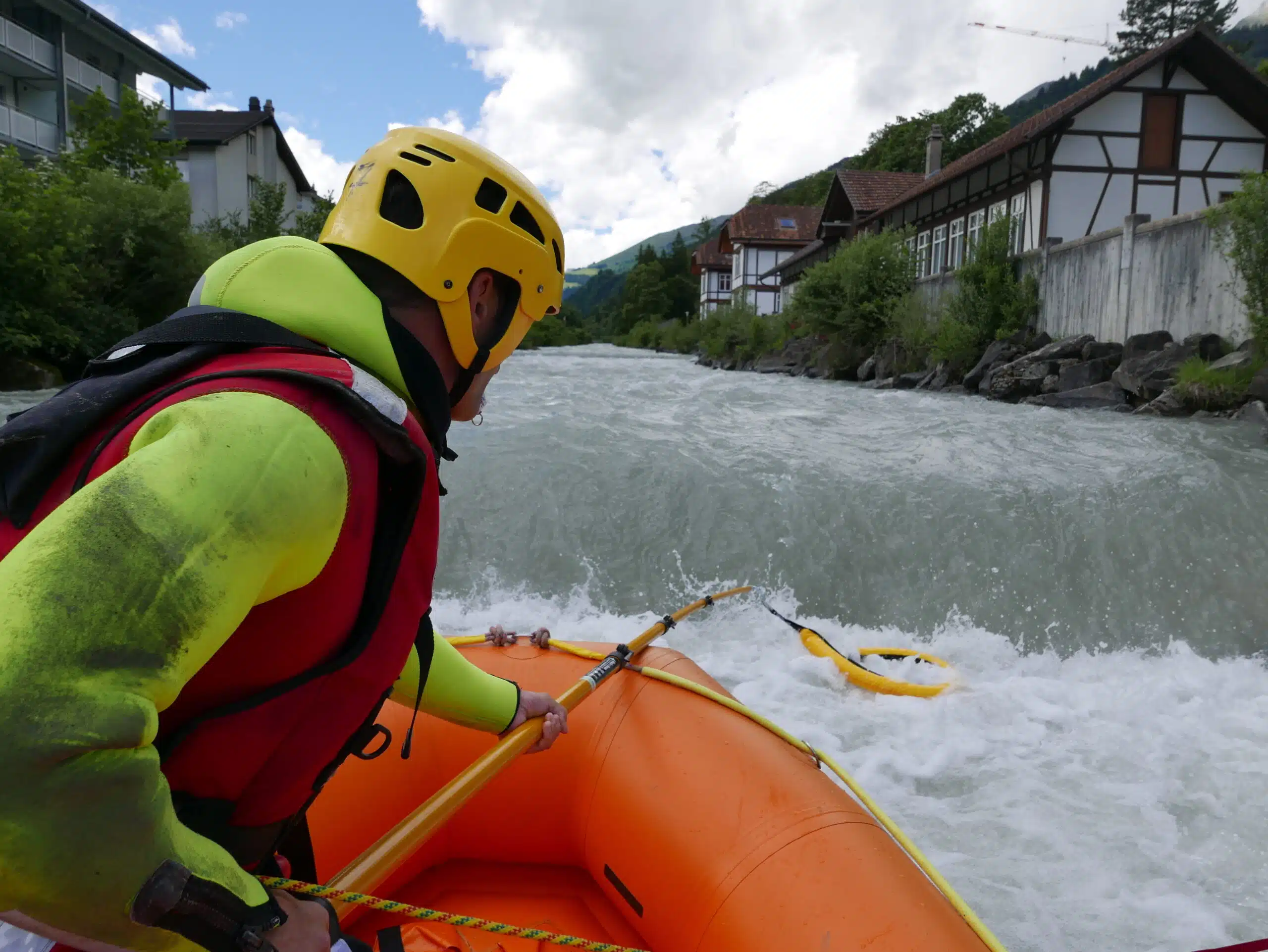 Soccorso Alluvionale e Fluviale Europa Persona con casco e giubbotto salvagente che guida un gommone arancione su un fiume in piena, superando sbarramenti artificiali e circondato da alberi e case. S.A.F.E.® - River Rescue