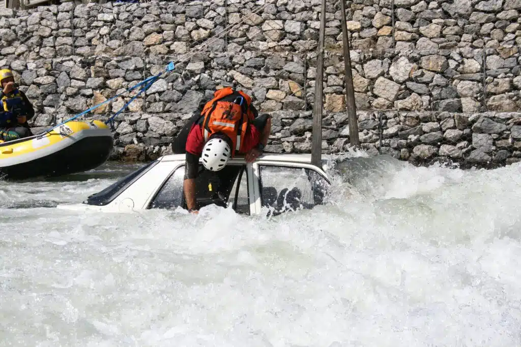 Soccorso Alluvionale e Fluviale Europa Un operatore del Rescue 3 è chino su un'auto sommersa in acque turbolente, vicino a un muro di pietra, con un gommone pronto all'azione. S.A.F.E.® - River Rescue