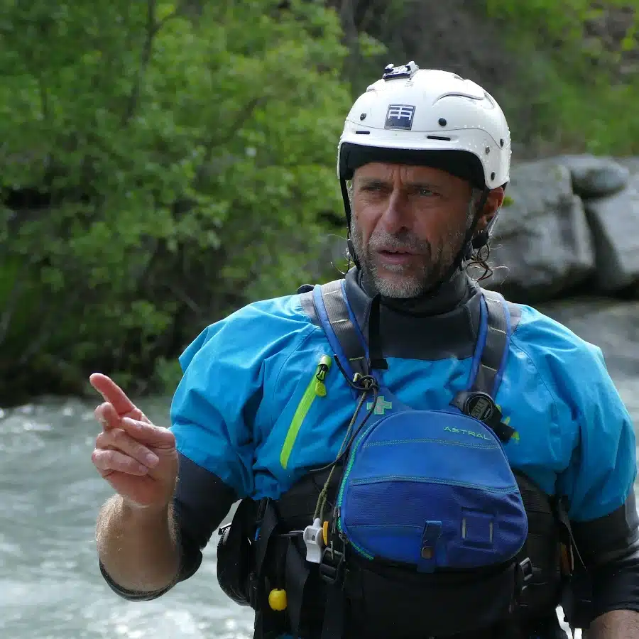 Soccorso Alluvionale e Fluviale Europa David Alemanni, che indossa un casco e un equipaggiamento blu, è in piedi sulla riva di un fiume, gesticolando con una mano, circondato da una vegetazione lussureggiante sullo sfondo. S.A.F.E.® - River Rescue