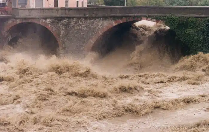 Soccorso Alluvionale e Fluviale Europa Le acque impetuose dell'alluvione si riversano sotto un ponte di pietra, mentre l'acqua fangosa dell'alluvione schizza violentemente attraverso gli archi, mentre le squadre di soccorso interagenzia monitorano la scena. S.A.F.E.® - River Rescue